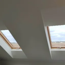 Dwa okna dachowe z drewnianą ramą w białym, skośnym suficie poddasza, widok na pole i pochmurne niebo.