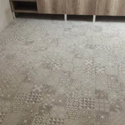 Ułożona podłoga z płytek ceramicznych o wzorze patchworkowym w odcieniach szarości i beżu, z widocznymi kartonami płytek przy ścianie i fragmentem szafki pod umywalkę.