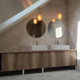 Ukończona łazienka na poddaszu ze skośnym sufitem, beżowe płytki, szafka z drewnopodobnym fornirem, dwie białe umywalki nablatowe, okrągłe lustra i wiszące lampy z żarówkami Edison.