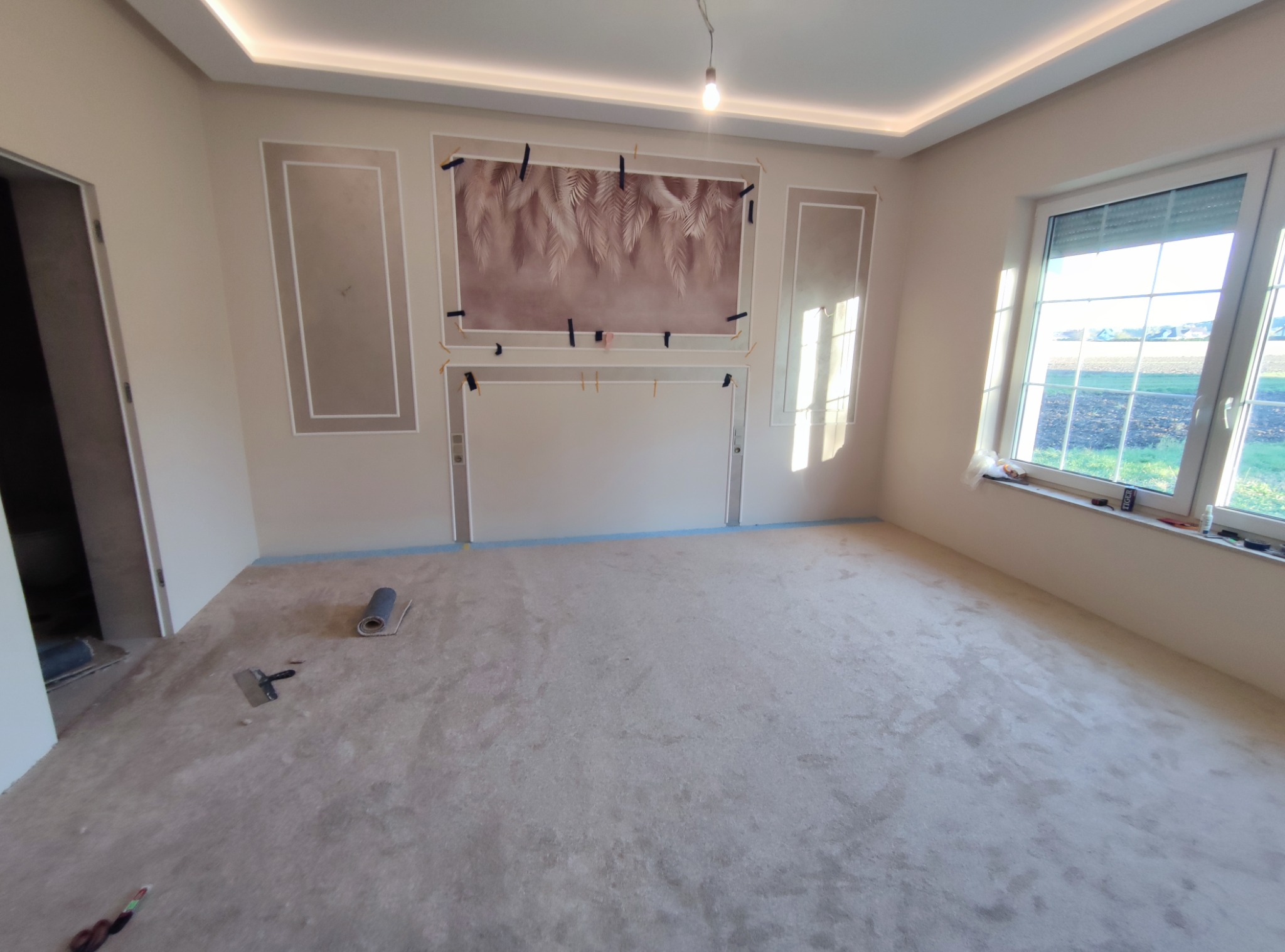 Pokój po remoncie: świeżo położona, jasna podłoga, ściany z dekoracyjnymi ramami i tapetą, okno z widokiem na zewnątrz. Widoczne narzędzia i materiały budowlane.