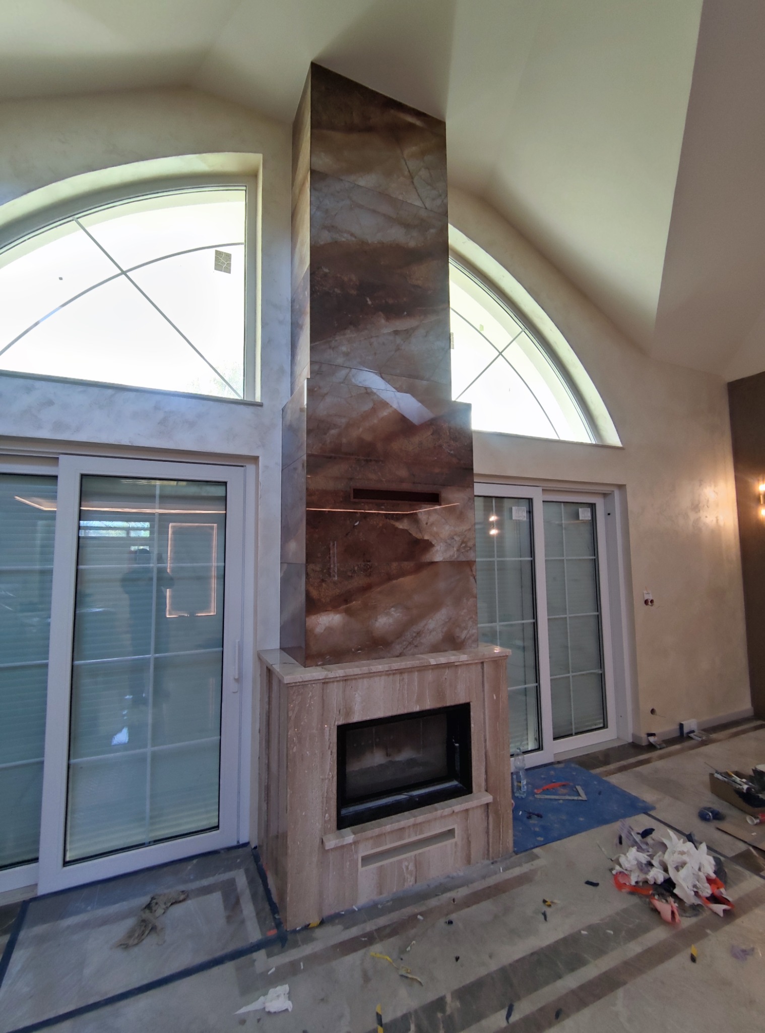 Wykończony kominek z okładziną z brązowego kamienia, z widocznym paleniskiem. W tle okna łukowe i przesuwne drzwi balkonowe. Podłoga w trakcie prac wykończeniowych.
