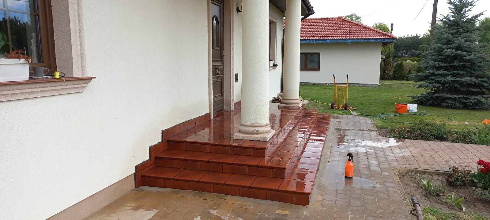 Wyłożone terakotą schody wejściowe do domu z kolumnami, po świeżym myciu. Widoczny fragment elewacji, kostka brukowa i narzędzia pracy.