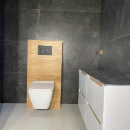 Nowoczesna łazienka z wiszącą miską WC na drewnianym panelu, ciemnymi płytkami na ścianach i białymi szafkami z drewnianymi wstawkami.