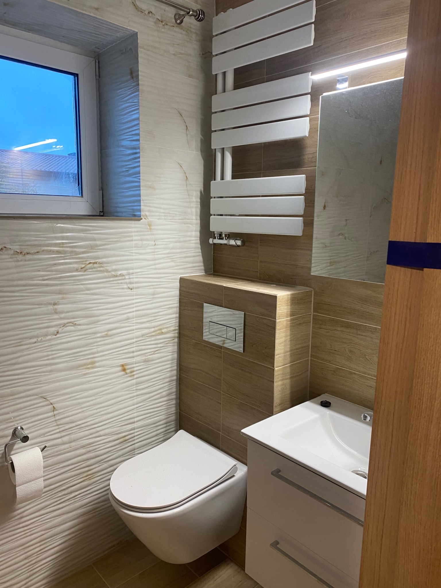 Nowoczesna łazienka z umywalką w szafce, podwieszaną toaletą i grzejnikiem drabinkowym, wykończona płytkami imitującymi marmur z falistą strukturą.