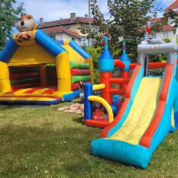 Kolorowy plac zabaw na trawie z dmuchanym zamkiem ze zjeżdżalnią i dmuchanym domkiem ze zwierzęciem na szczycie, w tle budynki mieszkalne.