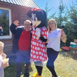 Osoba w stroju Myszki Minnie pozuje z dwoma osobami dorosłymi na tle drewnianego domku i zieleni, jedna z osób trzyma mikrofon.