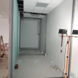 Montaż ścian działowych z płyt kartonowo-gipsowych w biurze, widoczne profile, płyty w kolorze zielonym, rozłożone narzędzia i oświetlenie budowlane.