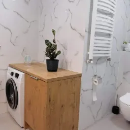 Nowoczesna łazienka z pralką zabudowaną w drewnianej szafce z blatem, marmurowe płytki na ścianach, grzejnik drabinkowy i roślina doniczkowa na blacie.