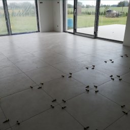 auto-mat Mateusz Ferenc - Układanie szarych płytek podłogowych w dużym, jasnym pomieszczeniu z widokiem na ogród przez duże okna; widoczne kliny i krzyżyki poziomujące płytki.