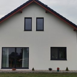 auto-mat Mateusz Ferenc - Elewacja domu jednorodzinnego w jasnym odcieniu z czarnymi ramami okiennymi, widoczne dwa okna na poddaszu i dwa na parterze, dach pokryty czarną dachówką z brązowymi elementami wykończeniowymi,...