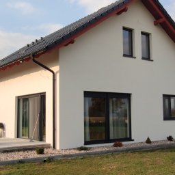 auto-mat Mateusz Ferenc - Nowoczesny dom jednorodzinny z białymi ścianami, czarną stolarką okienną i grafitową dachówką, widok z ogrodu na elewację boczną i taras z meblami ogrodowymi.