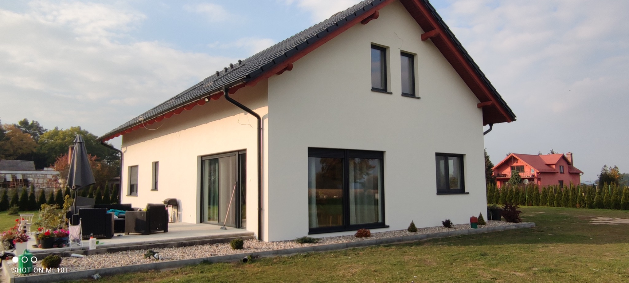 Nowoczesny dom jednorodzinny z białymi ścianami, czarną stolarką okienną i grafitową dachówką, widok z ogrodu na elewację boczną i taras z meblami ogrodowymi.