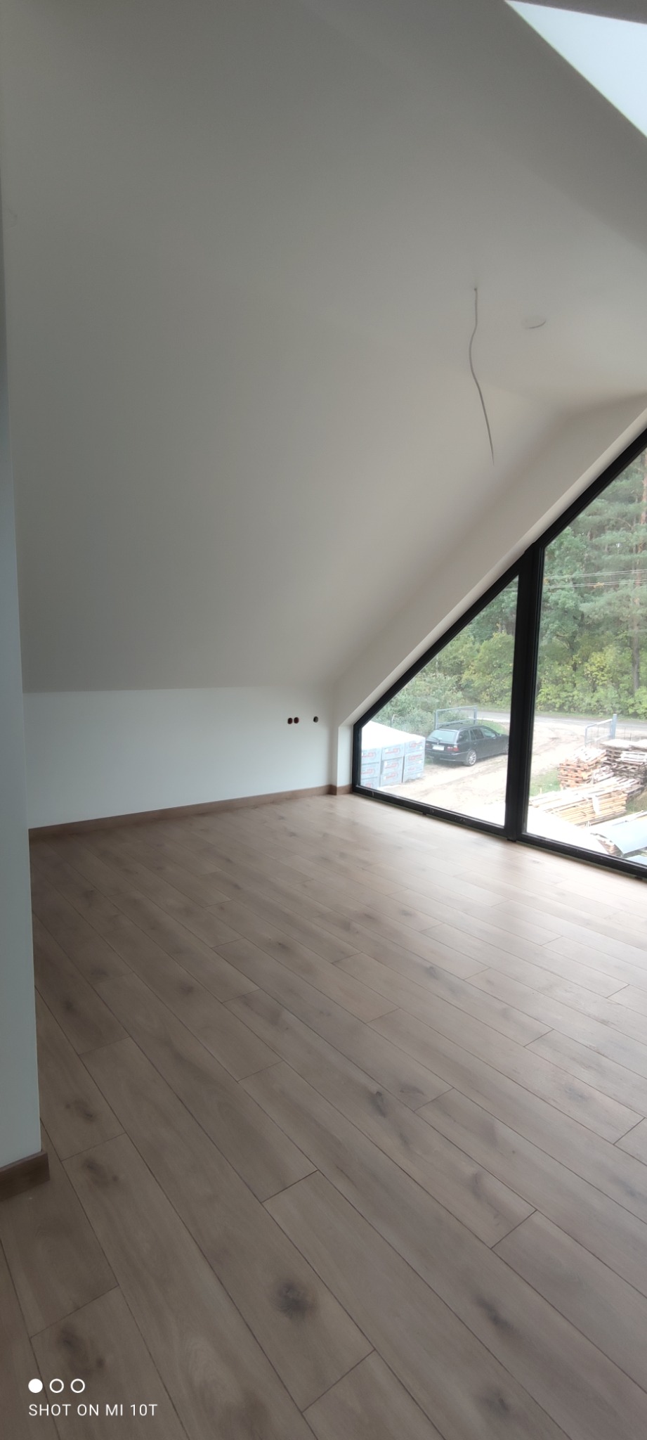 Poddasze z drewnianą podłogą i oknem w kształcie trójkąta, widok na zewnątrz z samochodem i materiałami budowlanymi.