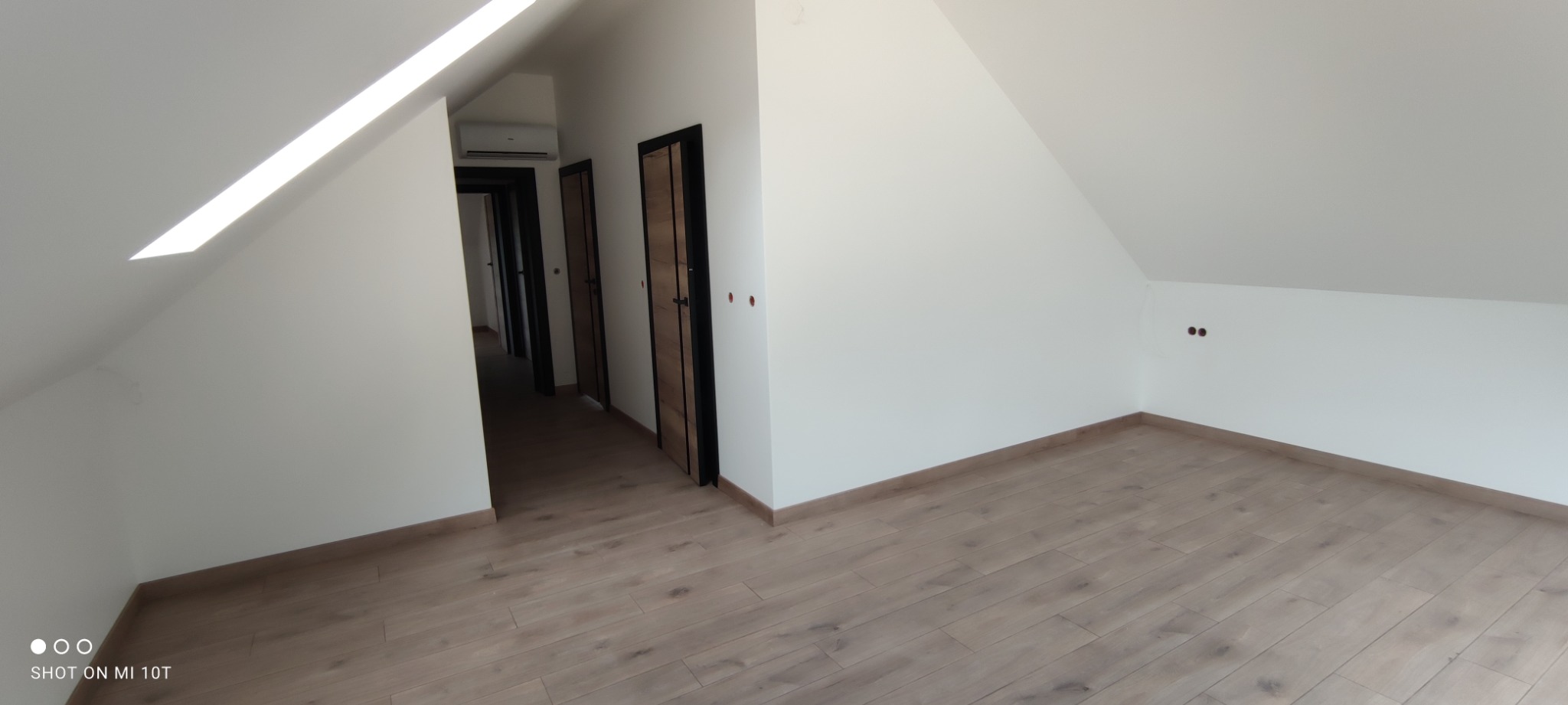 Poddasze z jasnymi panelami podłogowymi, białymi ścianami i trzema parami czarnych drzwi z drewnianym wypełnieniem w głębi, widoczne gniazdka elektryczne na ścianie.