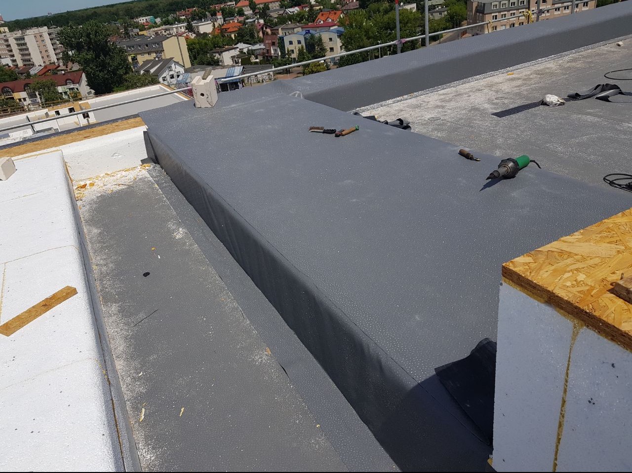 Szary dach płaski z widoczną hydroizolacją z papy termozgrzewalnej, narzędzia dekarskie i fragment ocieplenia z białego styropianu oraz płyty OSB, w tle zabudowa miejska.
