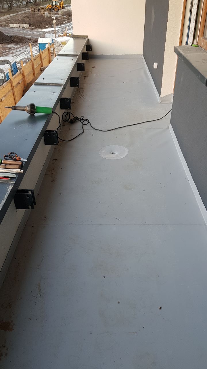 Szara, płynna izolacja balkonu w trakcie wykonywania, zgrzewarka na krawędzi, widoczne narzędzia i czarne wsporniki balustrady.
