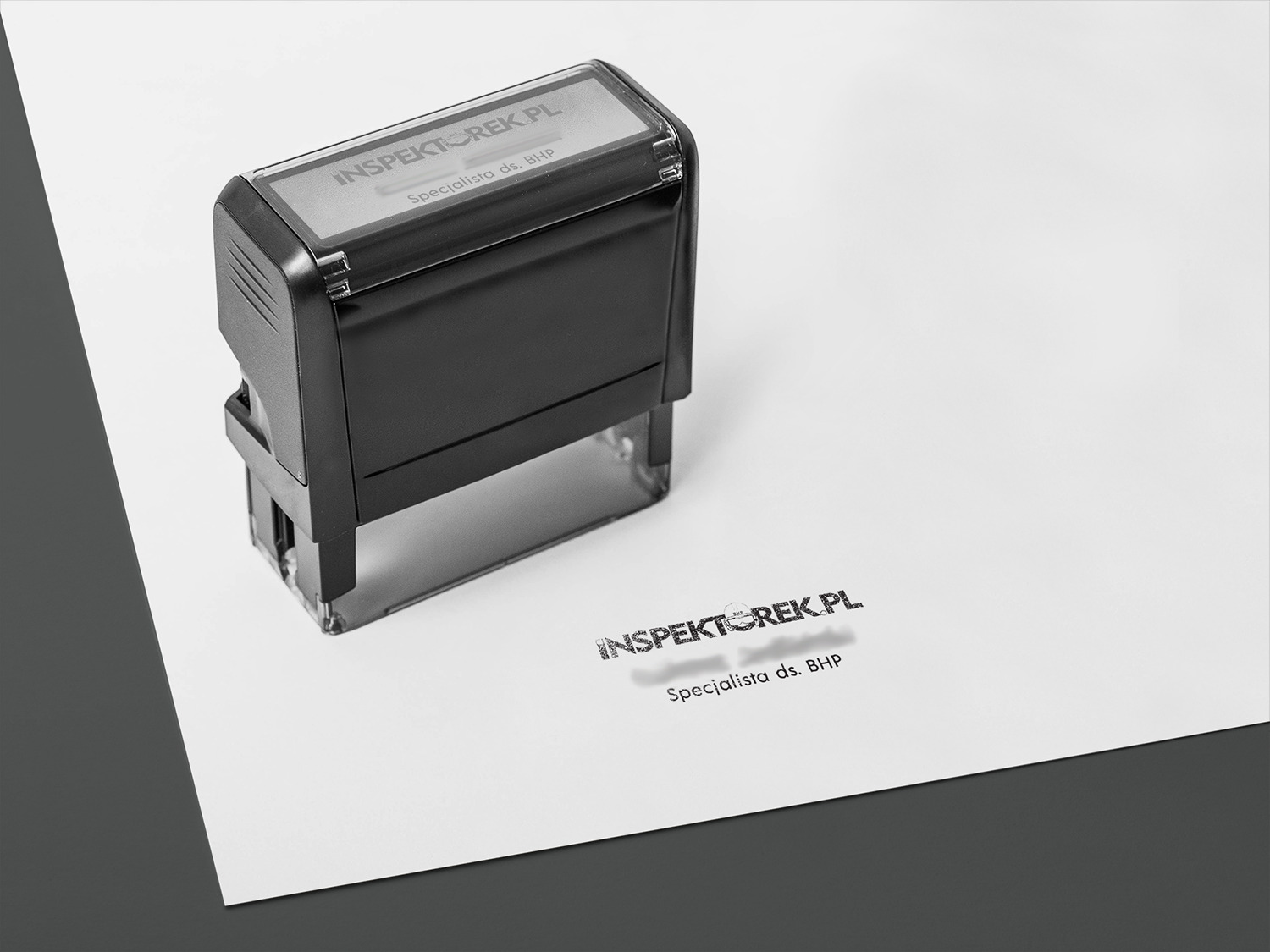 Czarna pieczątka automatyczna z napisem INSPEKTOREK.PL Specjalista ds. BHP odbita na białej kartce papieru, leżącej na szarym tle.