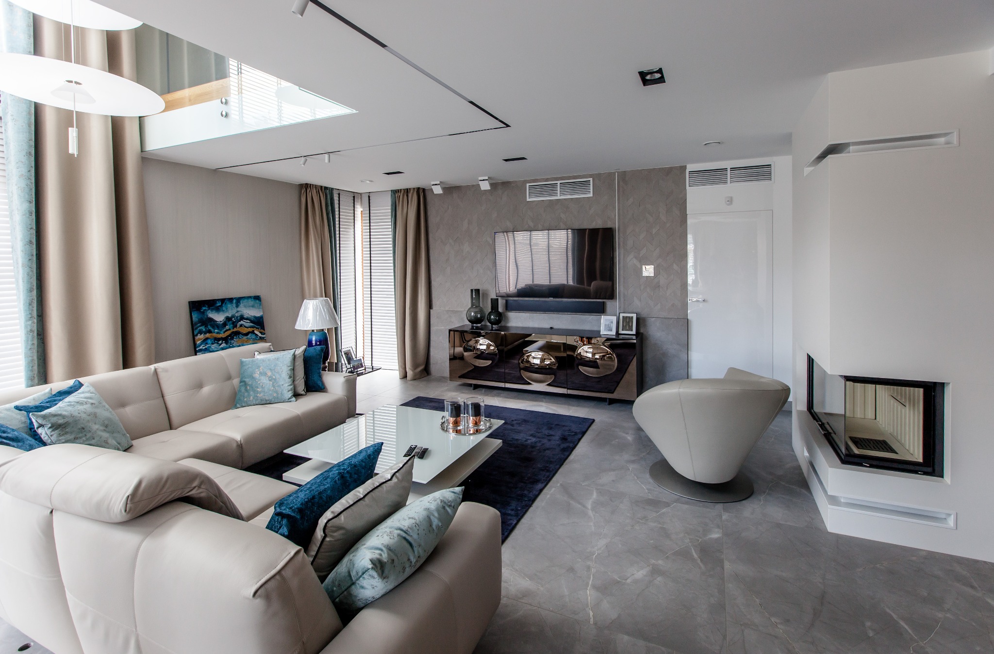 Jasny salon z narożną sofą, szklanym stolikiem kawowym na ciemnym dywanie, telewizorem na szafce z lustrzanymi frontami i nowoczesnym kominkiem w białej obudowie.