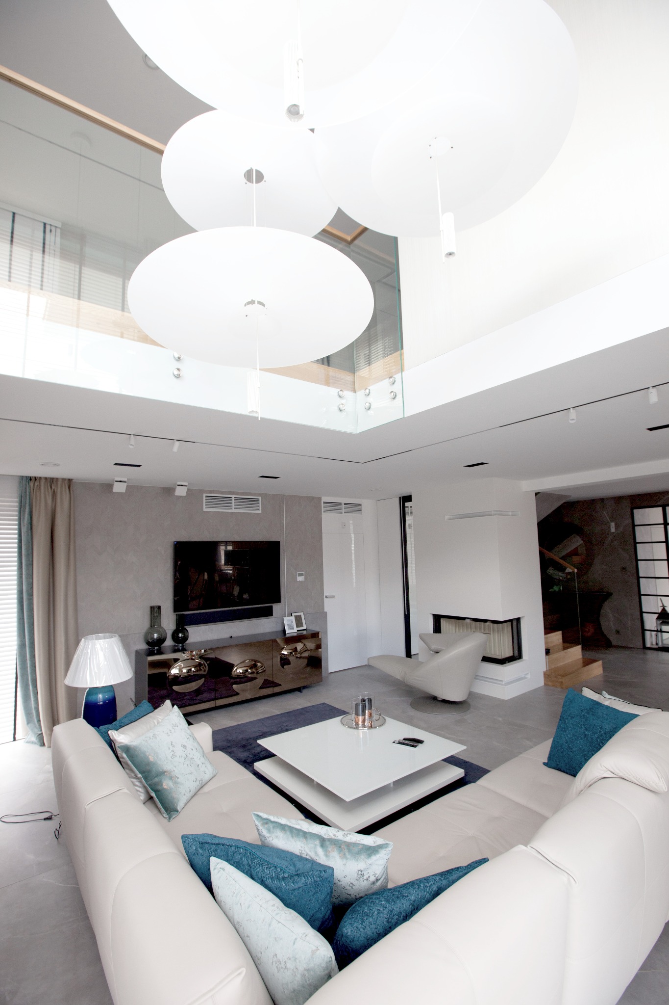 Przestronny salon w Lublinie z narożną, jasną sofą, turkusowymi poduszkami, nowoczesnym stolikiem kawowym i wiszącymi lampami w kształcie dysków, oświetlony naturalnym światłem z okna dachowego.