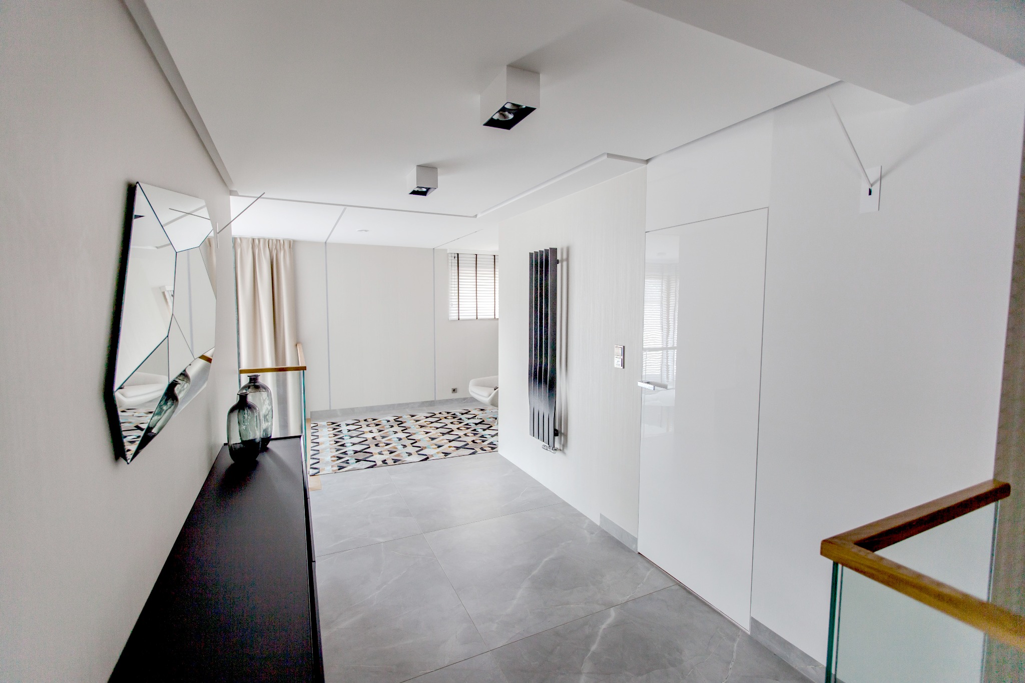 Nowoczesny, minimalistyczny przedpokój z szarą podłogą, czarną konsolą z dekoracyjnymi wazonami, geometrycznym lustrem i grzejnikiem żeberkowym.