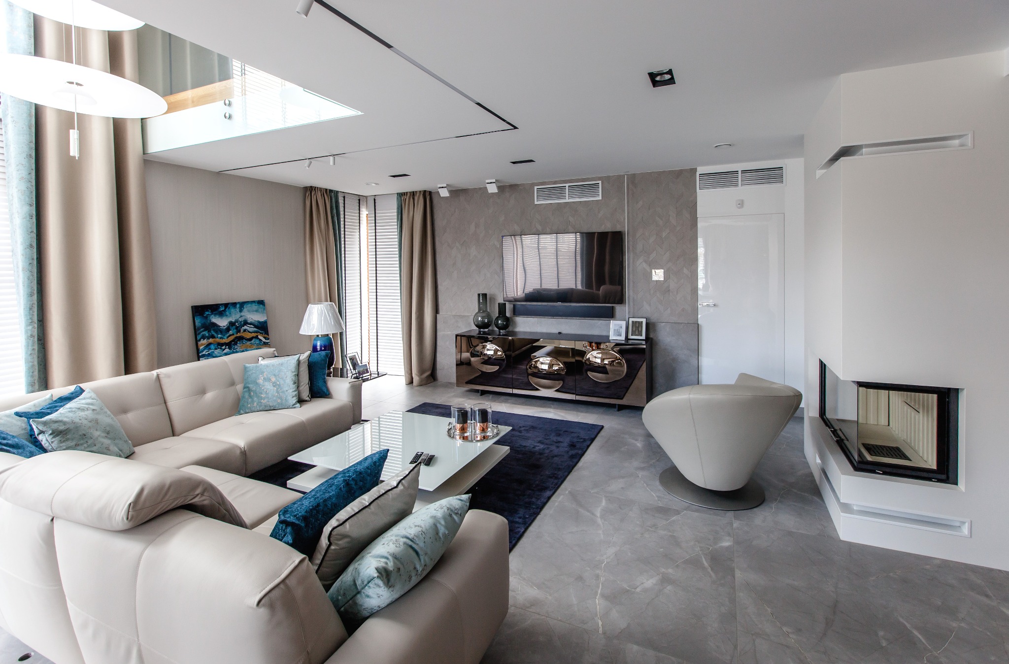 Nowoczesny salon z narożną sofą w odcieniach beżu i błękitu, szklanym stolikiem kawowym, ciemnym dywanem, szafką RTV z lustrzanymi frontami i kominkiem w zabudowie, oświetlony światłem dziennym...