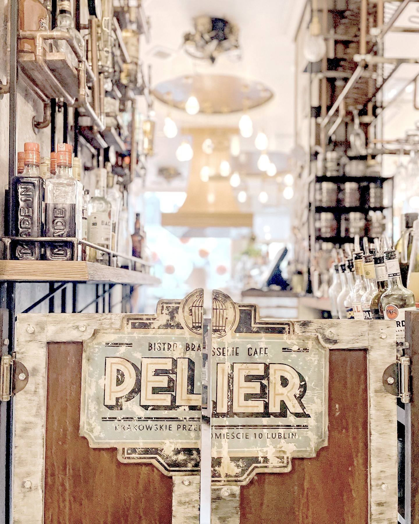 Wejście do baru bistro Pelier w Lublinie, z dekoracyjnymi drzwiami z logo, widok na półki z butelkami alkoholi i oświetlenie w tle.