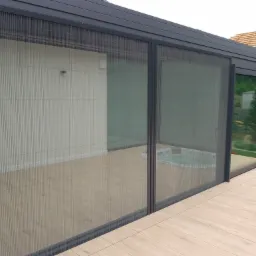 Szare, przesuwne panele z plisowanymi siatkami moskitierowymi, tworzące osłonę tarasu z widokiem na jacuzzi i ogród.