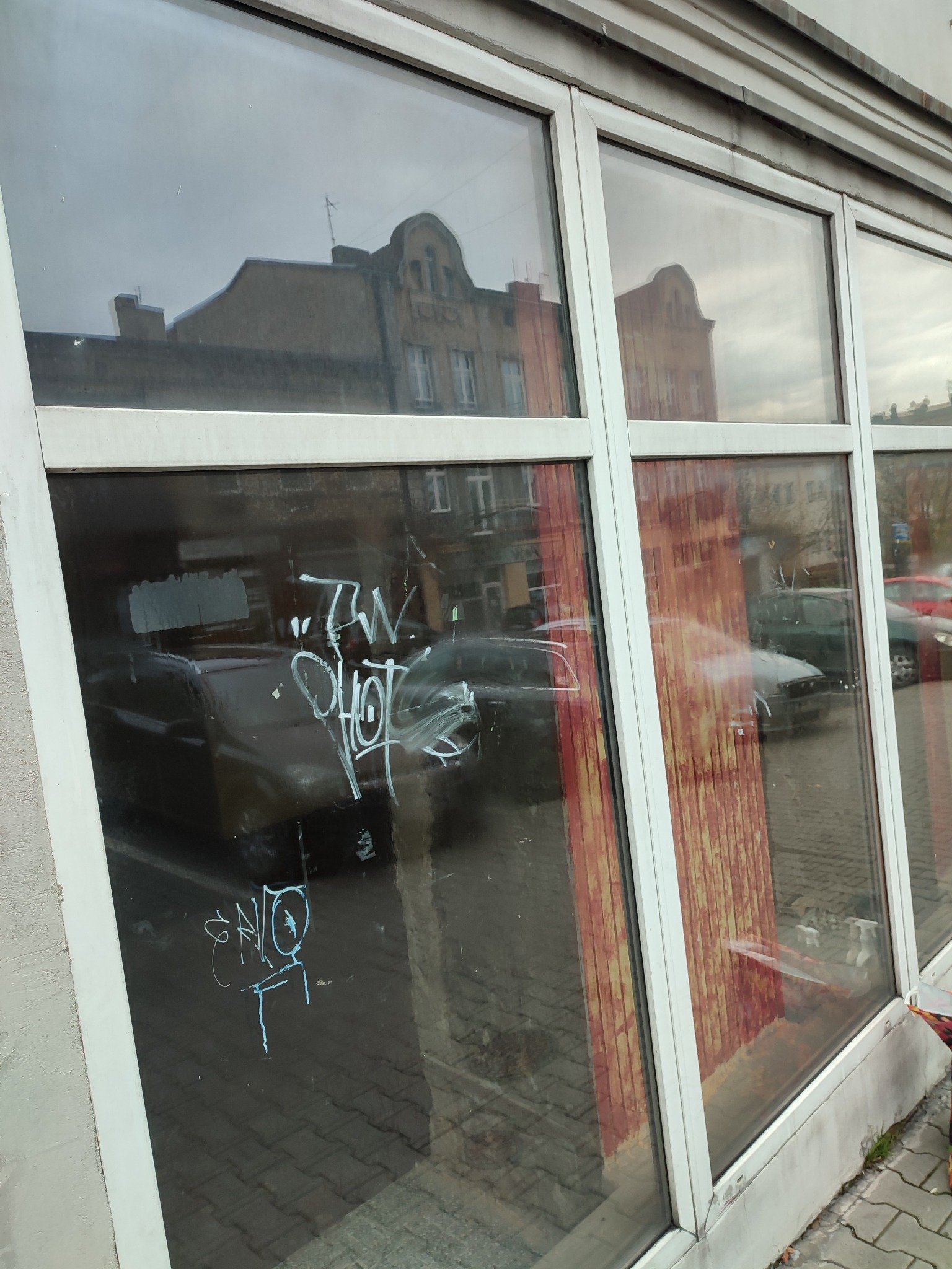 Okna witryny z białymi ramami zabrudzone graffiti, widoczne odbicia budynków i samochodów na ulicy.
