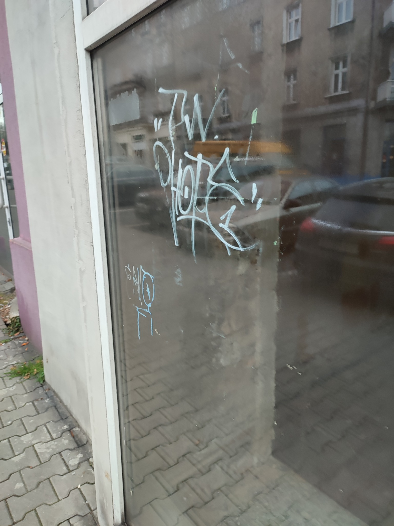 Zabrudzona szyba okienna z białymi napisami graffiti, widok z zewnątrz na chodnik z kostki i ulicę z zaparkowanymi samochodami, zarys budynku w tle.