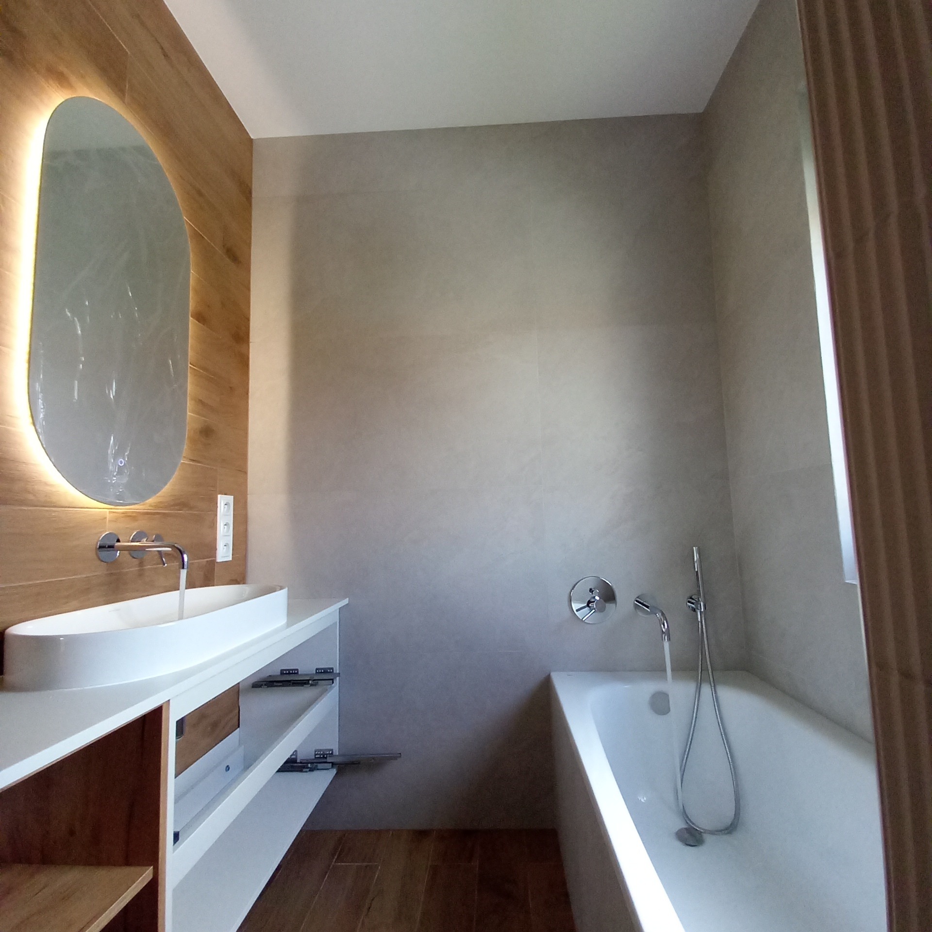 Nowoczesna łazienka z podświetlanym lustrem o organicznym kształcie, armaturą chromowaną, umywalką nablatową i wanną, wykończona płytkami imitującymi drewno i beton.