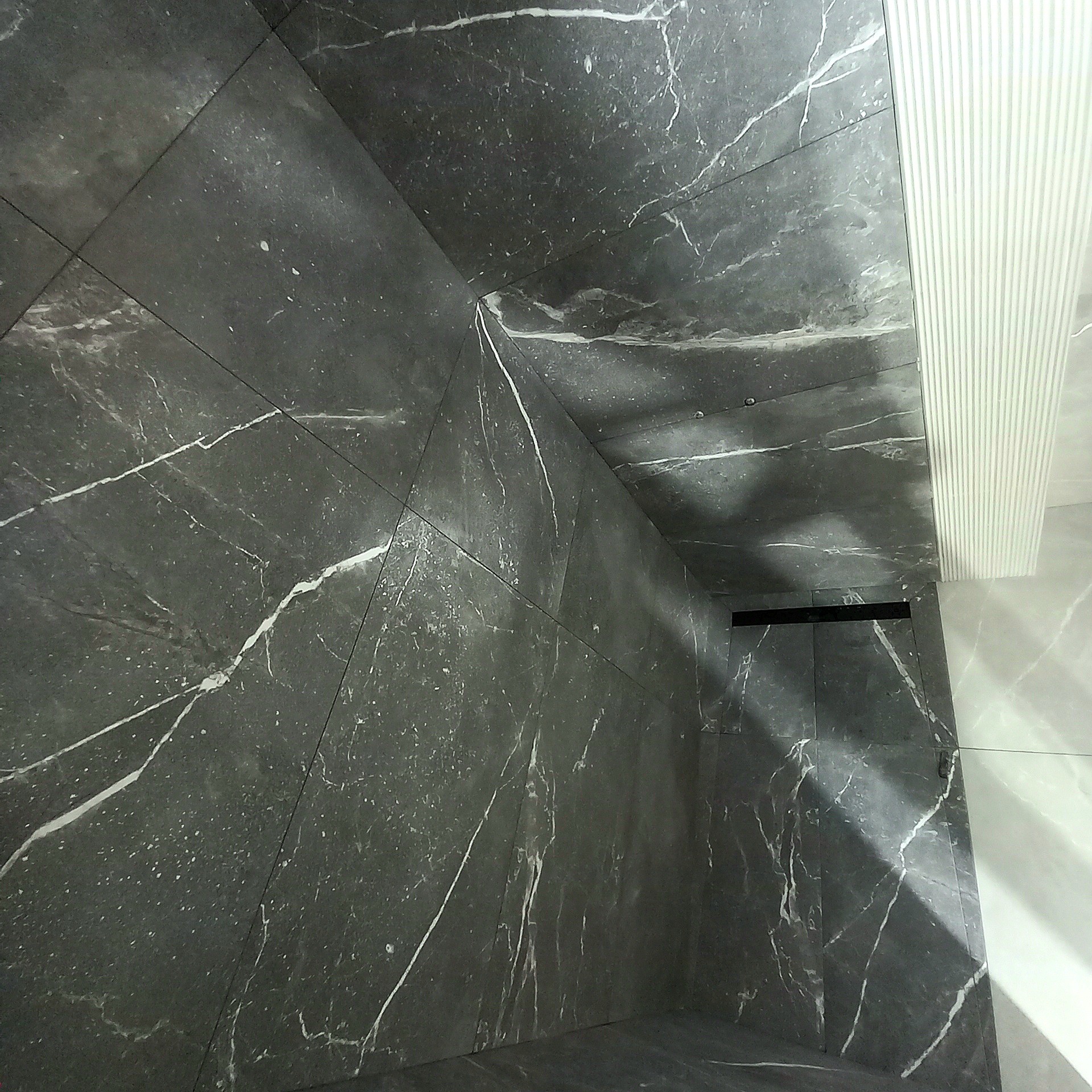 Wnętrze łazienki wykończone ciemnoszarymi płytkami imitującymi marmur z białymi żyłami, widoczny odpływ liniowy i fragment ściany z białymi, pionowymi panelami dekoracyjnymi.