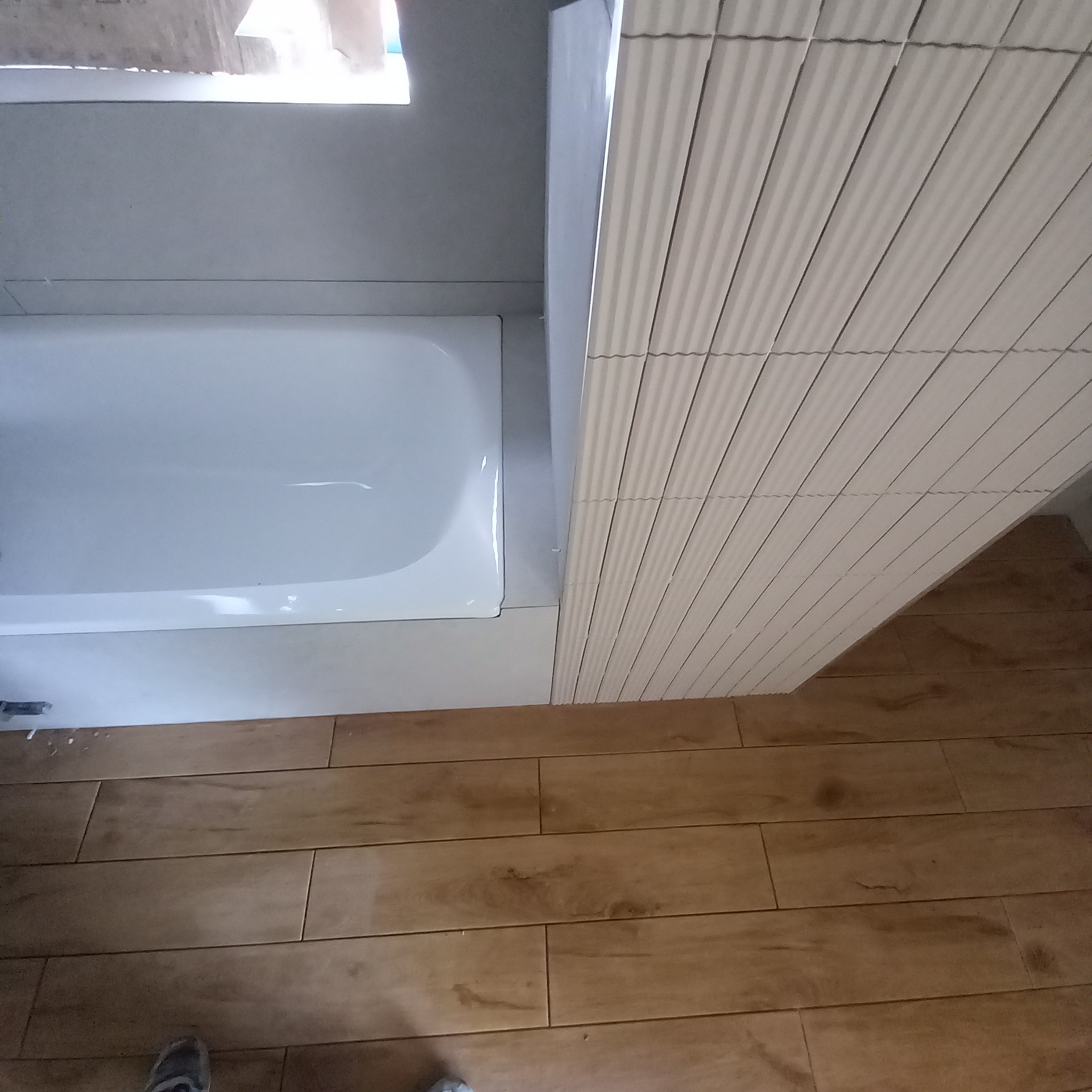 Wykończona łazienka z białą wanną, okładziną z płytek imitujących pionowe fale i podłogą z płytek drewnopodobnych ułożonych w cegiełkę.