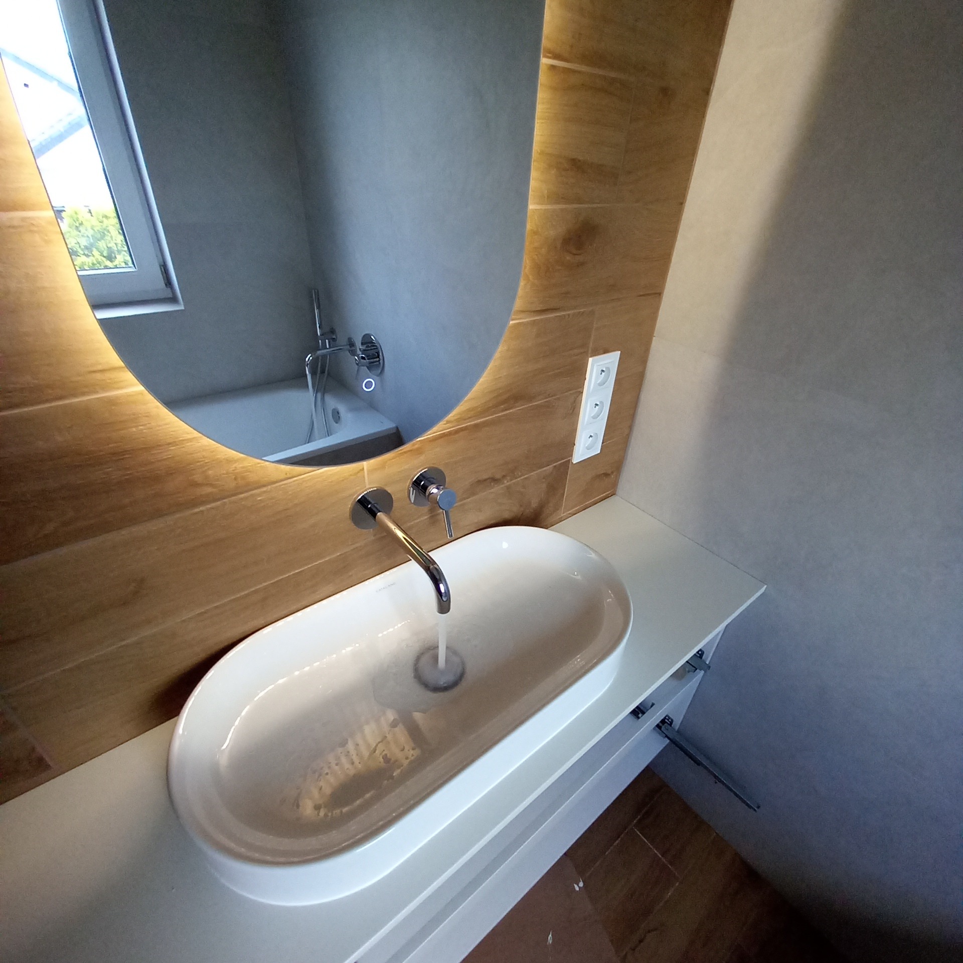 Nowoczesna łazienka z owalną umywalką nablatową, chromowaną baterią i podświetlanym lustrem w drewnianej ramie, widoczne gniazdka elektryczne i fragment wanny odbijającej się w lustrze.