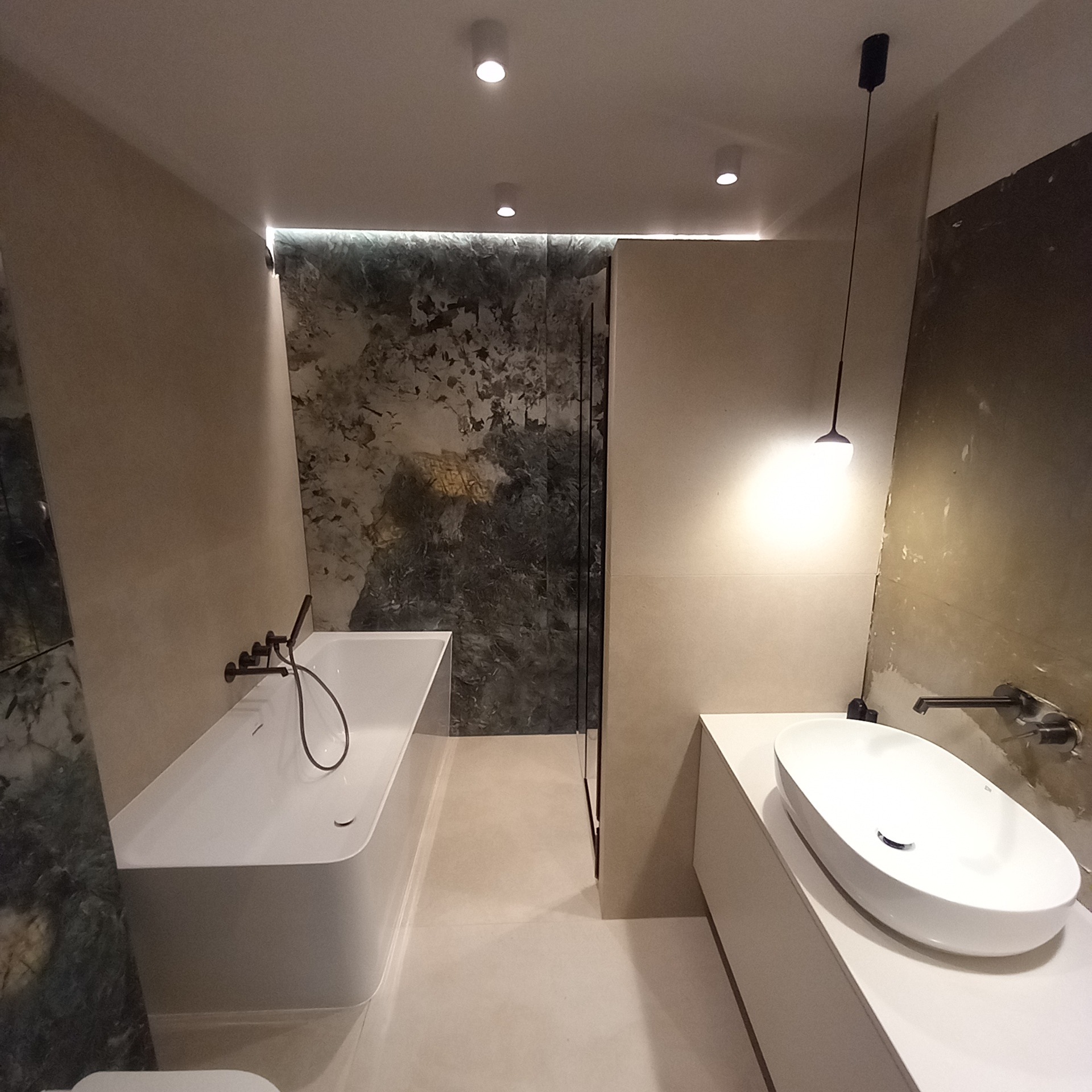 Nowoczesna łazienka z białym blatem, owalną umywalką, czarną armaturą i ścianą wykończoną płytami imitującymi kamień z wyraźnym wzorem, oświetlona punktowymi lampami sufitowymi i wiszącą lampą.