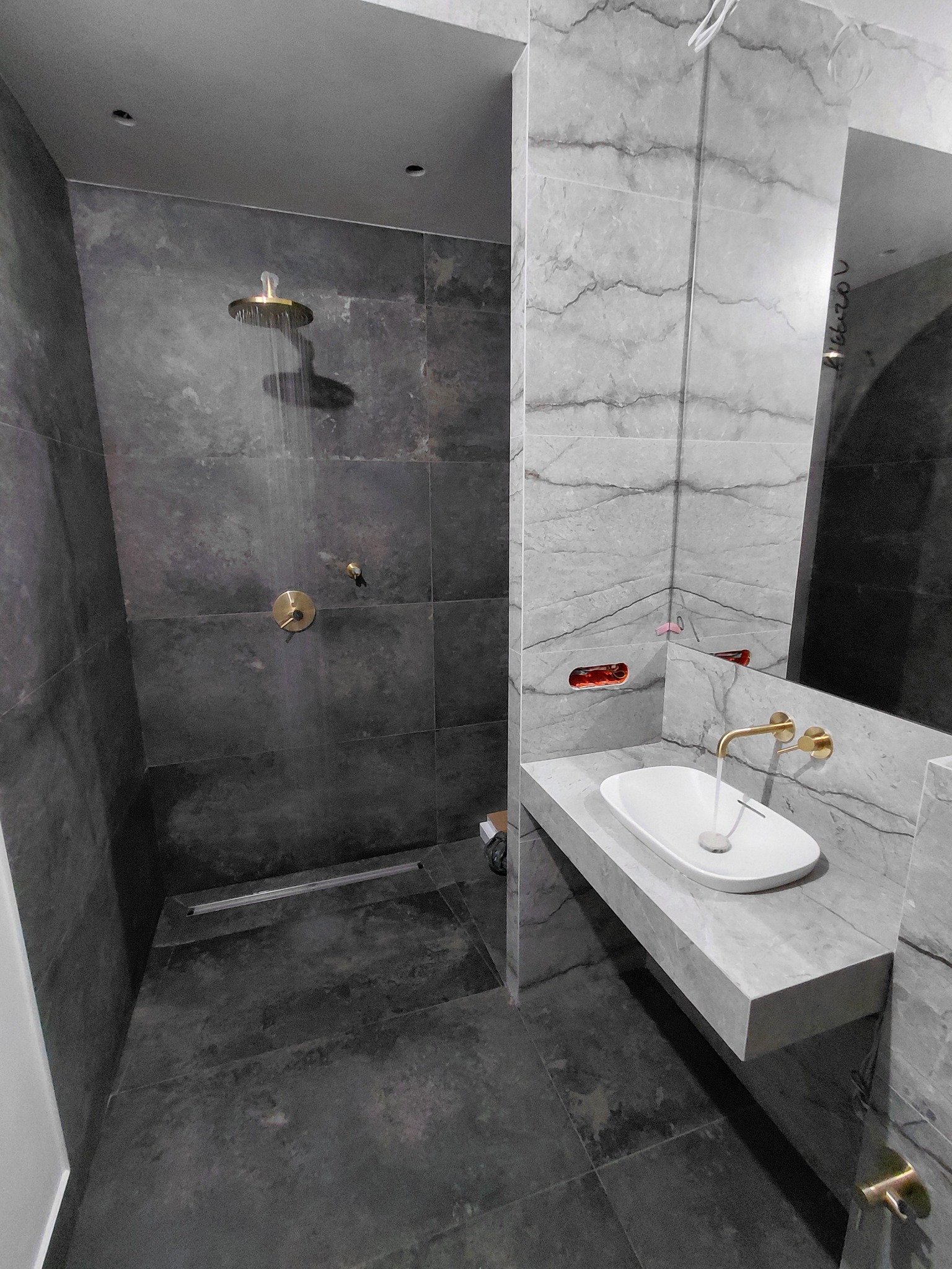 Nowoczesna łazienka z marmurowymi płytkami, złota armatura, deszczownica w użyciu. Minimalistyczny design z podwieszaną umywalką i odpływem liniowym.