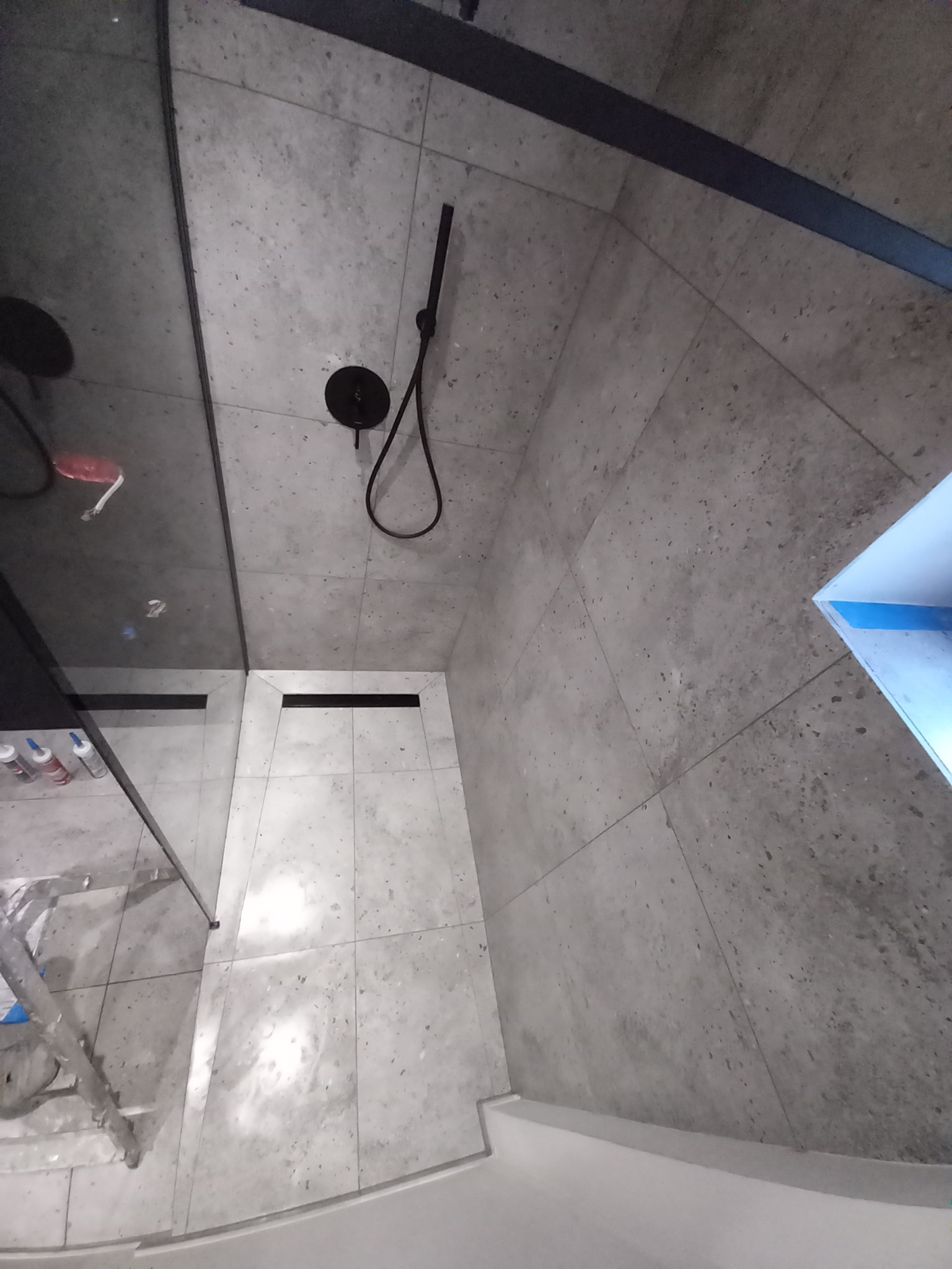 Wykończona łazienka z szarymi płytkami, czarną armaturą i odpływem liniowym. Widoczny fragment drabiny malarskiej i akcesoria. Ujęcie pod kątem.