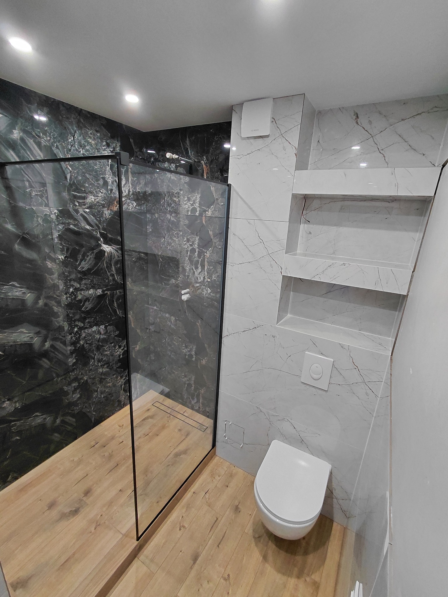 Nowoczesna łazienka po remoncie: kabina typu walk-in z czarnymi profilami, drewnopodobna podłoga, marmurowe płytki na ścianach i wnęki półkowe.