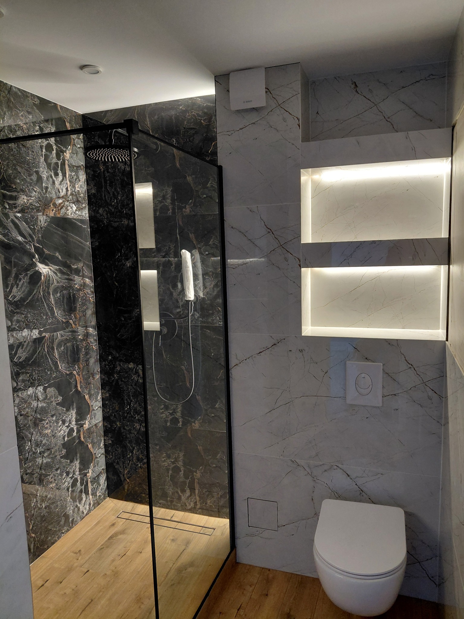 Nowoczesna łazienka z kabiną typu walk-in, wykończona marmurowymi płytkami w odcieniach szarości i czerni. Drewnopodobna podłoga i podświetlane wnęki dodają elegancji.