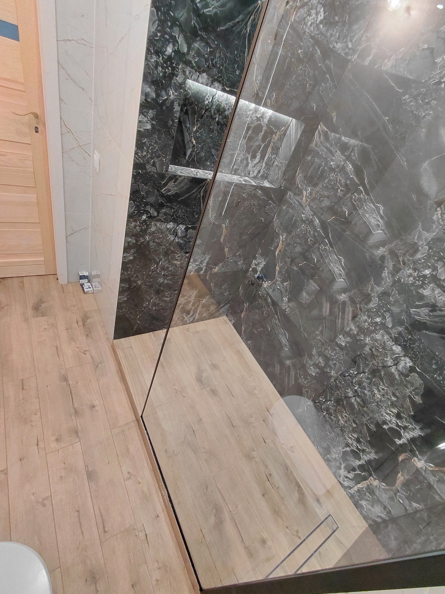 Nowoczesna łazienka z kabiną typu walk-in, wykończona płytkami imitującymi marmur w odcieniach szarości i beżu. Widoczne detale wykończenia i drewnopodobna podłoga.