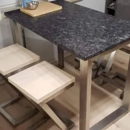 Stół barowy z blatem z ciemnego granitu i metalową konstrukcją oraz dwa drewniane hokery z metalową podstawą w nowoczesnej kuchni.