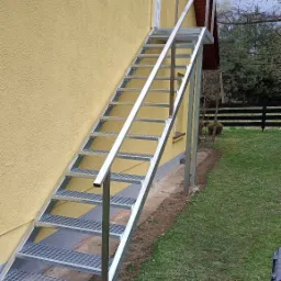 Zewnętrzne metalowe schody z kratowymi stopniami i prostą poręczą prowadzące do drzwi na piętrze żółtego budynku.