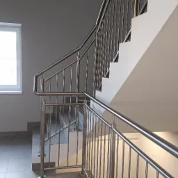 Wewnętrzne schody z metalowymi poręczami i balustradami o minimalistycznym designie, widoczne z dołu, z szarymi płytkami na podłodze i oknem w tle.