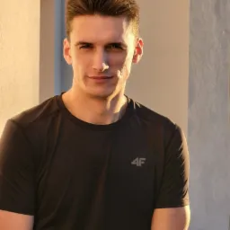 Portret mężczyzny w czarnej koszulce sportowej z logo 4F, oświetlonego ciepłym światłem, na tle jasnej ściany w Krakowie.