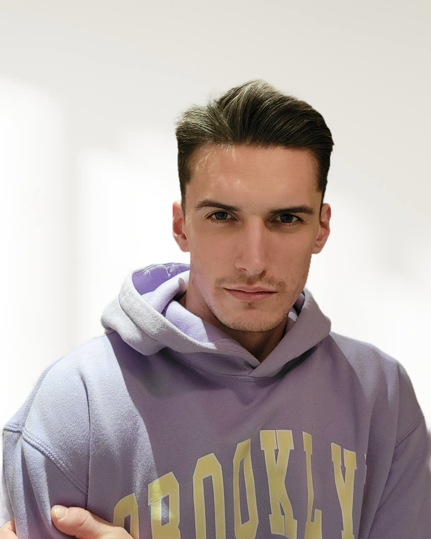 Portret młodego mężczyzny z zaczesanymi do tyłu włosami, ubranego w fioletową bluzę z kapturem z napisem BROOKLYN.