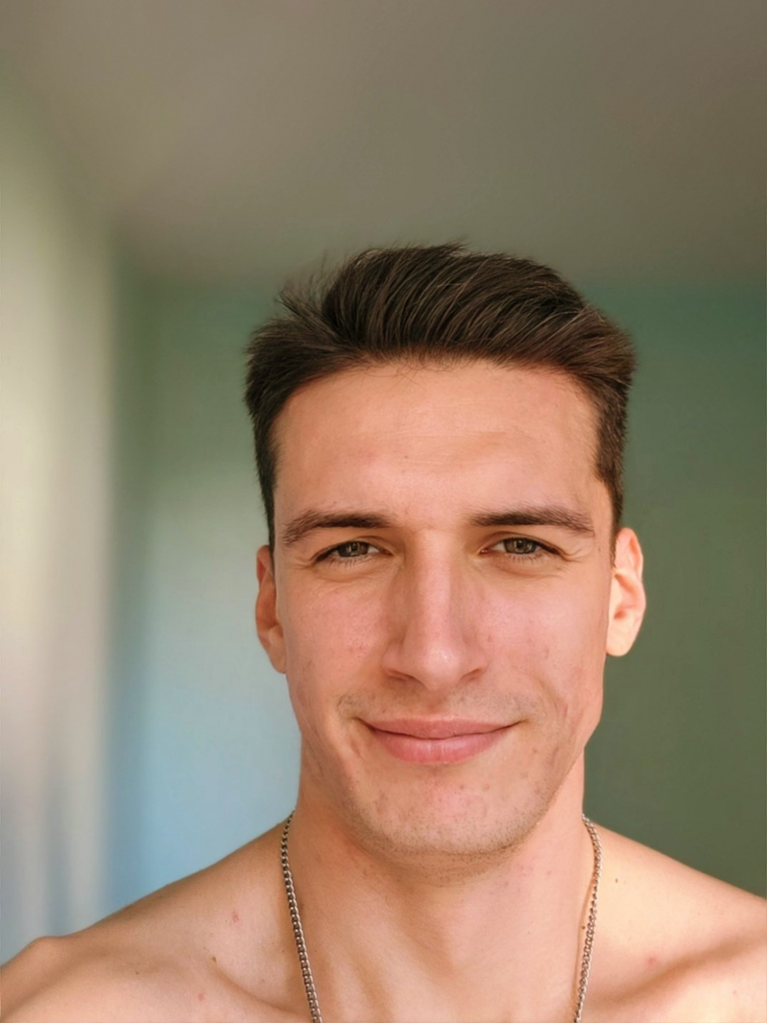 Portret uśmiechniętego młodego mężczyzny z krótkimi, ciemnymi włosami i srebrnym łańcuszkiem na szyi, na rozmytym tle w odcieniach zieleni i beżu.