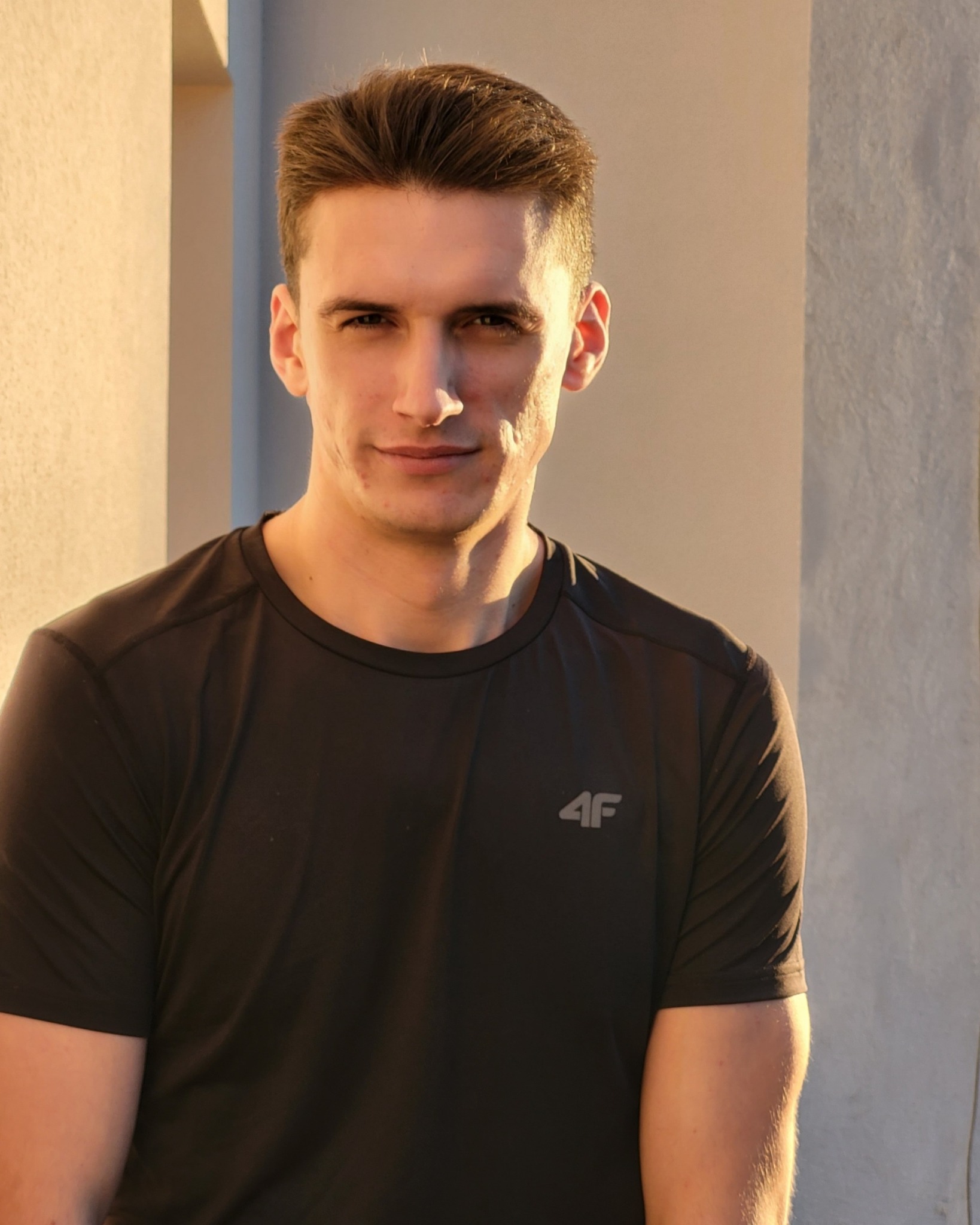 Portret mężczyzny w czarnej koszulce sportowej z logo 4F, oświetlonego ciepłym światłem, na tle jasnej ściany w Krakowie.
