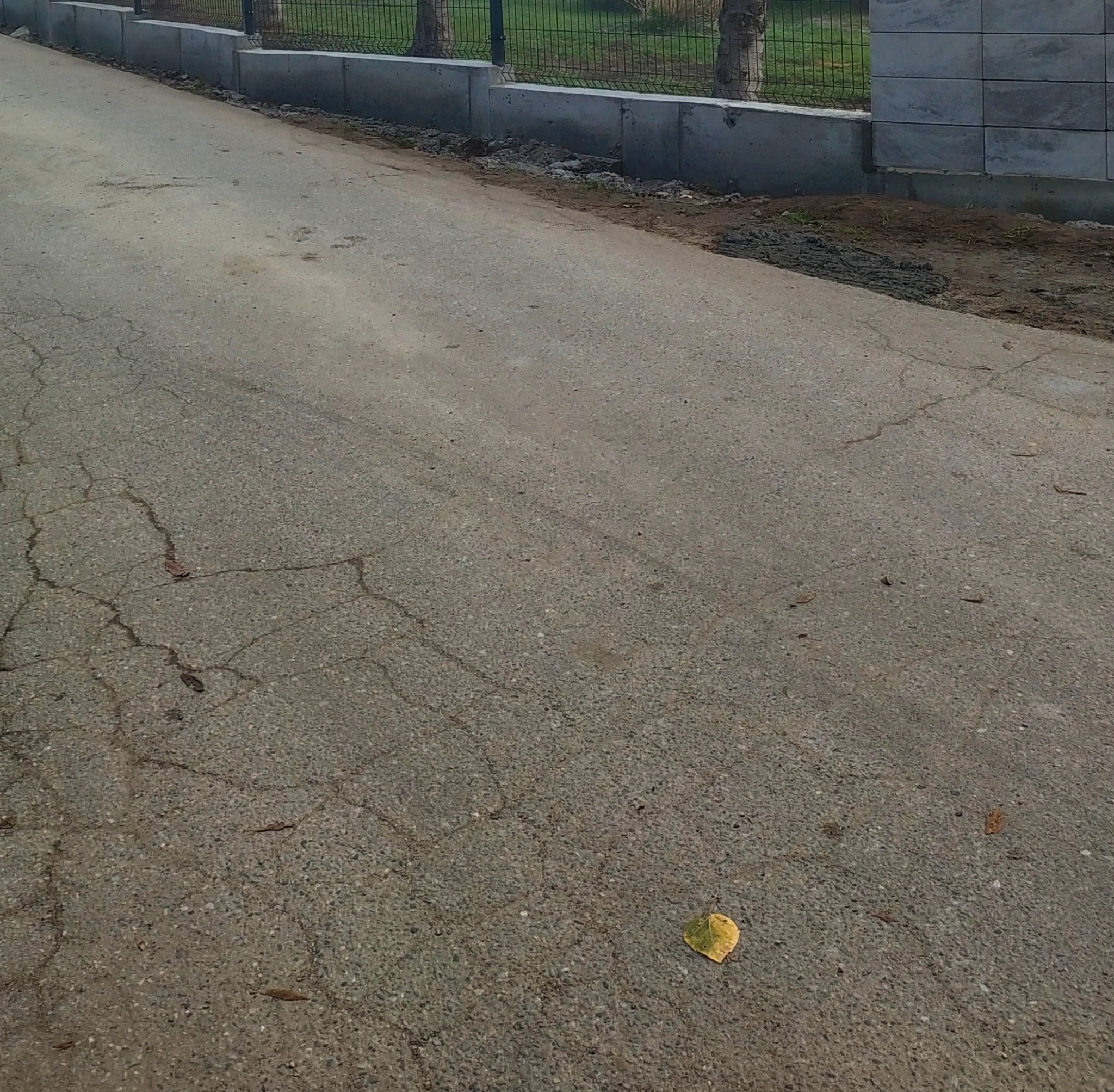Asfaltowa droga z pęknięciami, obok nowy betonowy murek z metalową siatką ogrodzeniową i pojedynczy żółty liść na asfalcie.