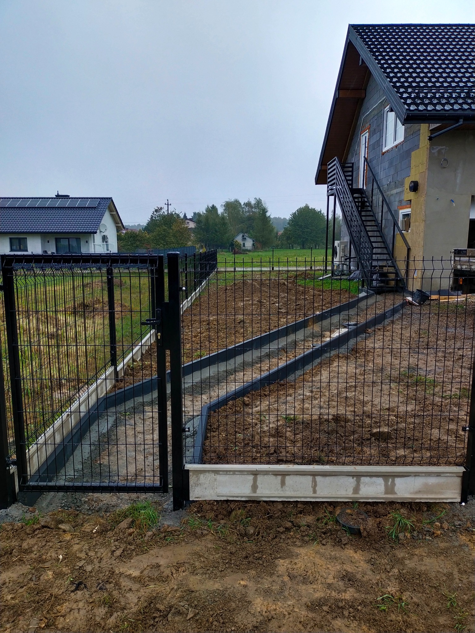 Czarna metalowa siatka ogrodzeniowa z otwartą bramą, widoczne betonowe obrzeża i przygotowany teren pod nasadzenia, w tle dom w trakcie budowy z zewnętrznymi schodami.
