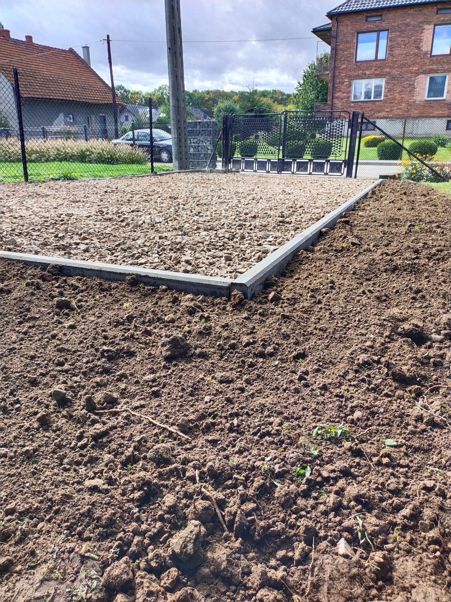 Przygotowany podkład z kamienia i obrzeży pod przyszłą nawierzchnię, widoczna brama wjazdowa i ogrodzenie, w tle dom jednorodzinny.