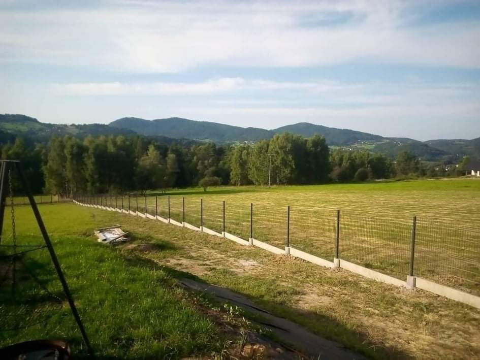 Długa siatka ogrodzeniowa z betonową podmurówką, biegnąca wzdłuż rozległego, zielonego pola z widokiem na zalesione wzgórza w tle.